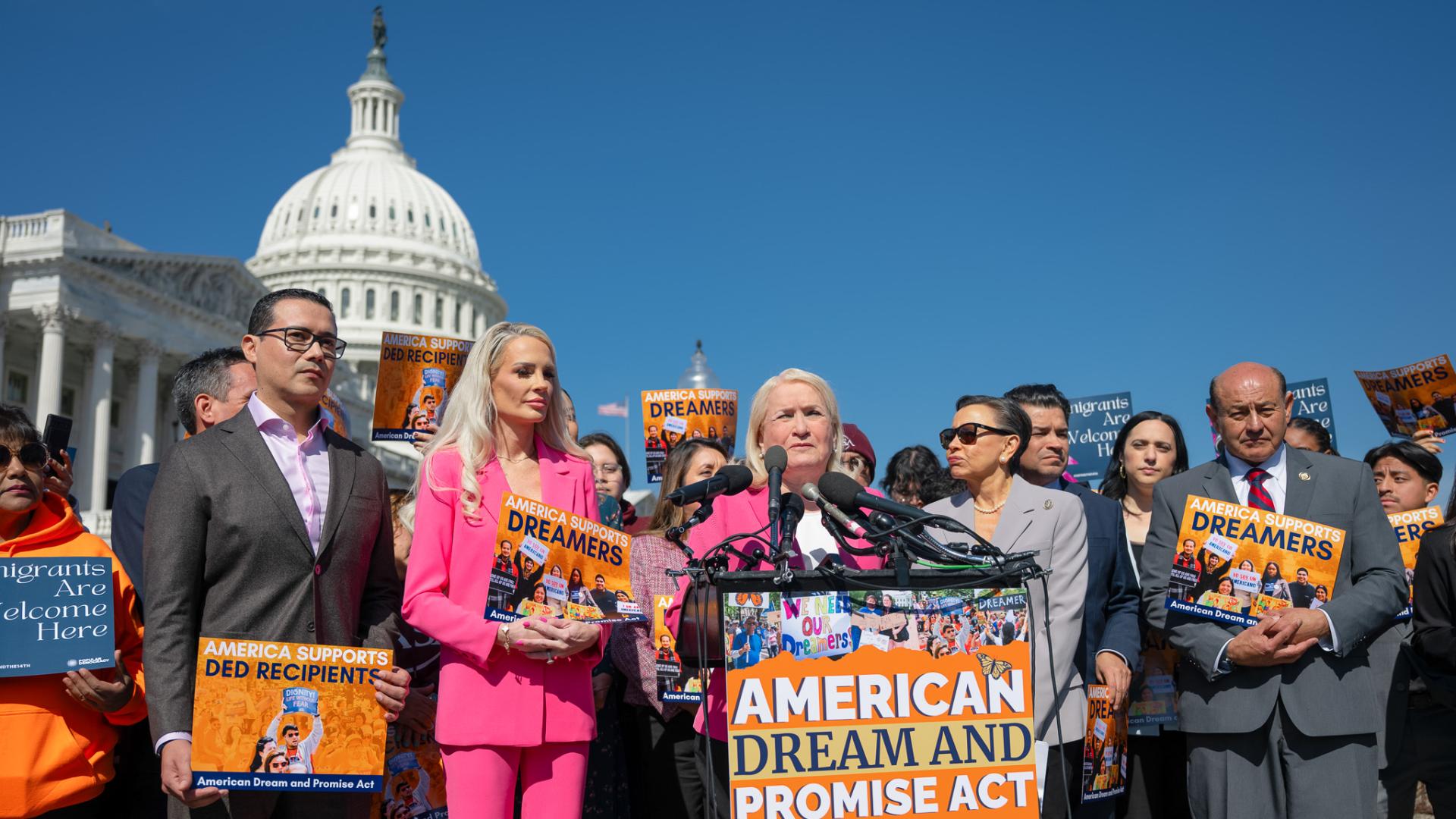 This image captures a press conference in support of Dreamers held in front of the U.S. Capitol. A group of politicians and advocates stand together, holding signs that read "America Supports Dreamers" and "America Supports DED Recipients." The event appears to be focused on immigration policy, particularly the American Dream and Promise Act.  At the podium, Rep. Sylvia Garcia in a pink blazer speaks into multiple microphones, with a determined expression.